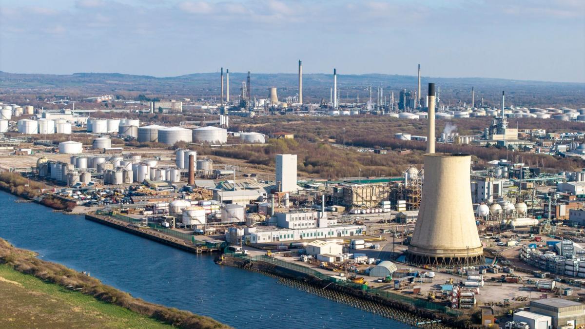 The Manchester Shipping Canal can be seen running across the image, on the left bank is grass and on the right a large industrial complex stretching into the distance. There are rolling hills outlined in the background and a blue sky with some white clouds.