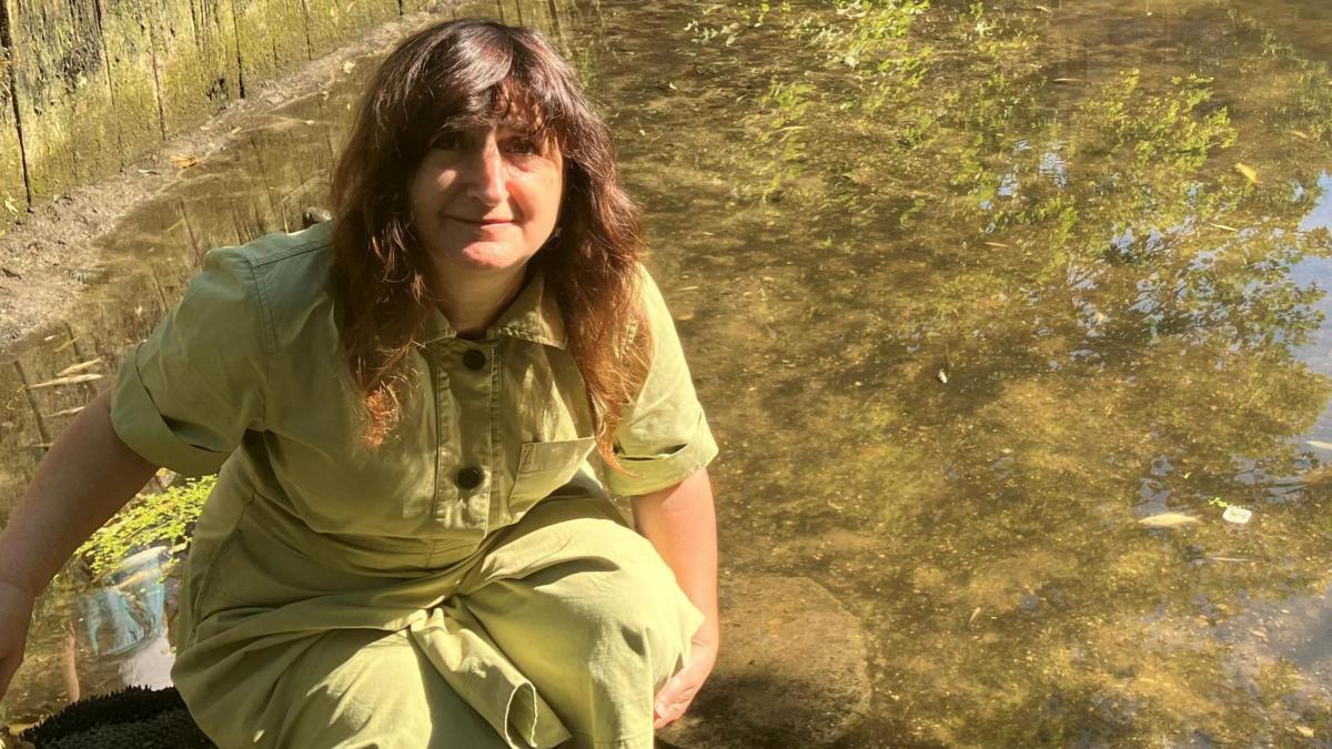 A woman wearing a light green jumpsuit kneeling down by the side of the water. She has wavy brown hair and is smiling while looking up to the camera. She is kneeling next to the river.