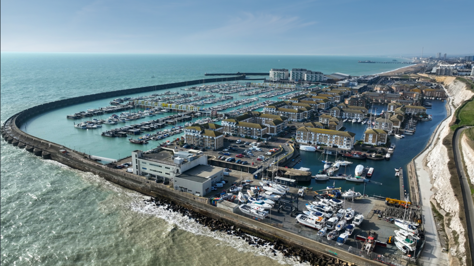 An aerial view of Brighton Marina shows curved harbour walls enclosing multiple pontoons filled with boats and yachts. Apartment blocks, marina buildings, roads and parking areas surround the basin. The sea extends into the distance alongside the Brighton coastline, with chalk cliffs visible on the