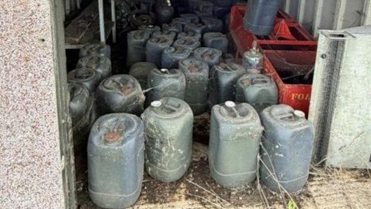 A number of dark green containers inside what appears to be an open building. There are two red crates on one side. The building has grey walls and there appears to be an earth floor.