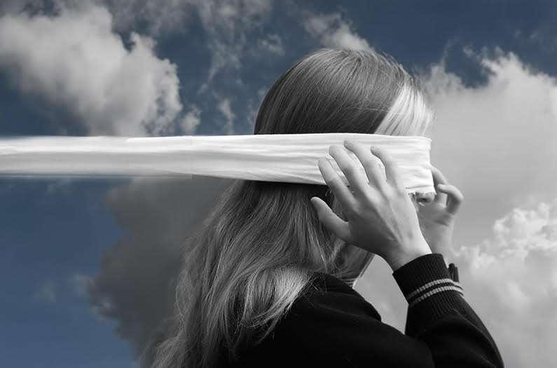 Person places a white blindfold over their eyes, seen from behind against a cloudy blue sky.