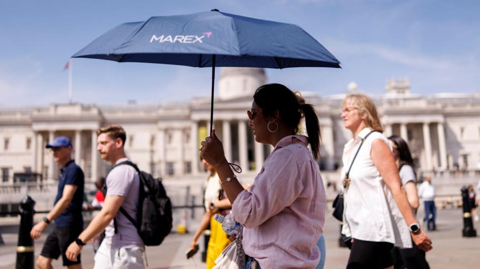 A woman uses an umbrella to shelter from the sun, surrounded by several other people, in a sun-drenched Trafalgar Square in London