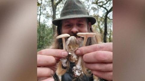 Colin Unsworth, wearing a dark outdoor hat, holds three olive amanita mushrooms, Amanita submembranacea, close to the camera with both hands. Two of the mushrooms have their pale gills visible beneath flat caps, while the smaller central mushroom has a rounded brownish cap. Woodland trees can be see