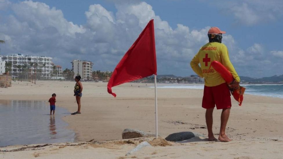 A lifeguard stands next to a red flag warning of hazardous sea conditions as Hurricane Polo approaches Baja California Sur in San Jose del Cabo, Mexico, on 26 September, 2026. The lifeguard is wearing a bright yellow long-sleeved shirt with the words "salvavidas" (Spanish for lifeguard) and "lifegua