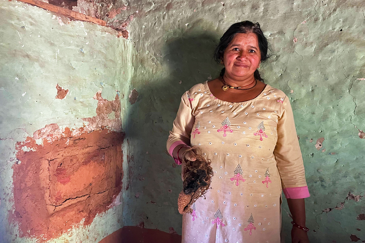 Kamla Devi stands beside the back cavity of her wall hive, sealed with a mixture of red soil and cow dung. She holds an old sack, which she burns to smoke out the bees during honeycomb harvest. — Image by Swati Thapa.