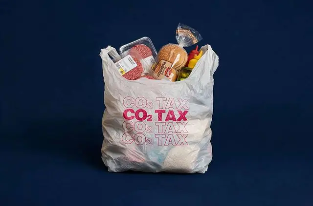 Grocery bag full of items against a dark blue background, with bold 'CO2 TAX' text on the bag, conveying a carbon tax theme.