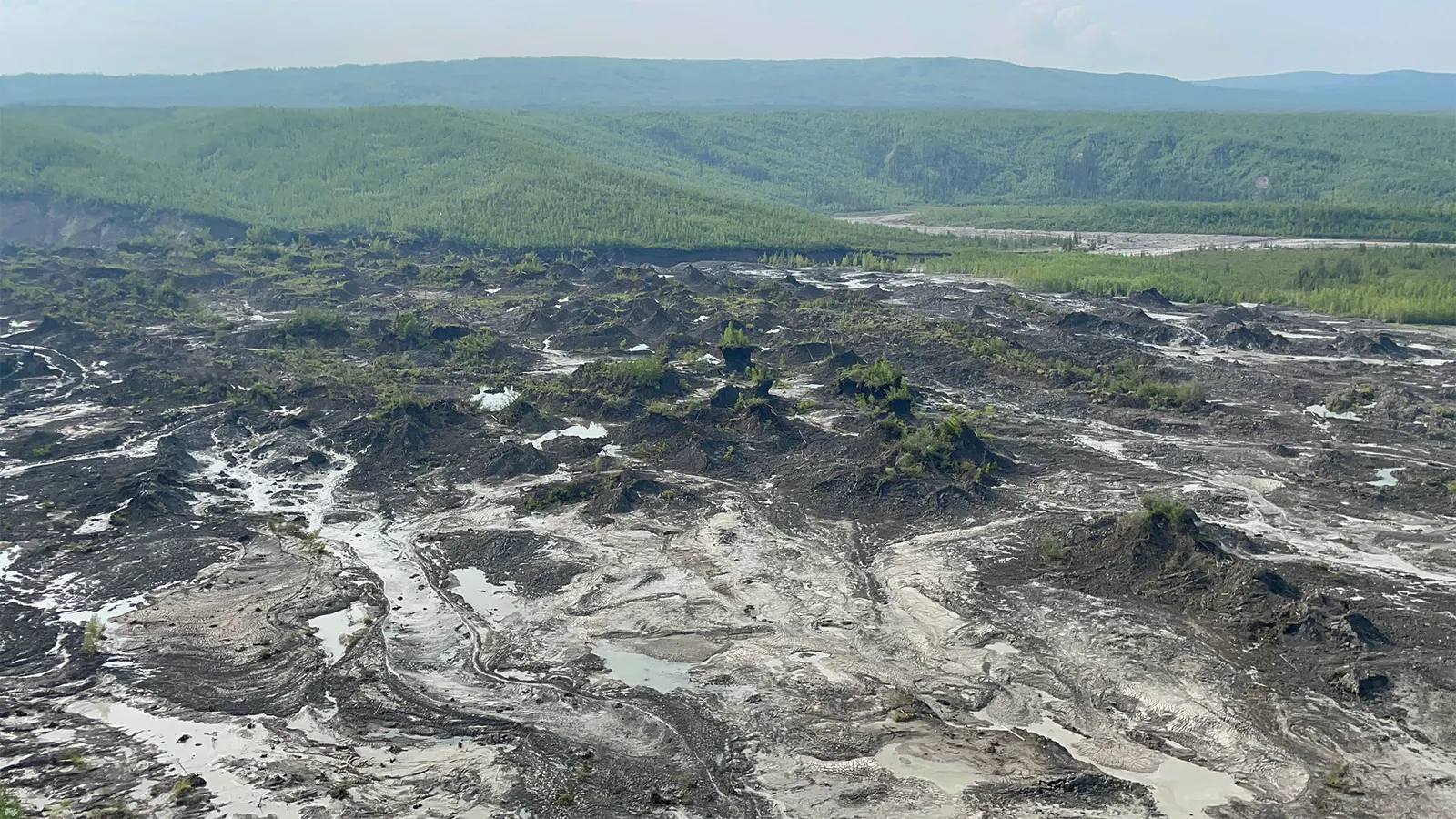 Image via Grist landscape showing damaged earth caused by permafrost detachment slides with tree covered hills in the background — Image via Grist