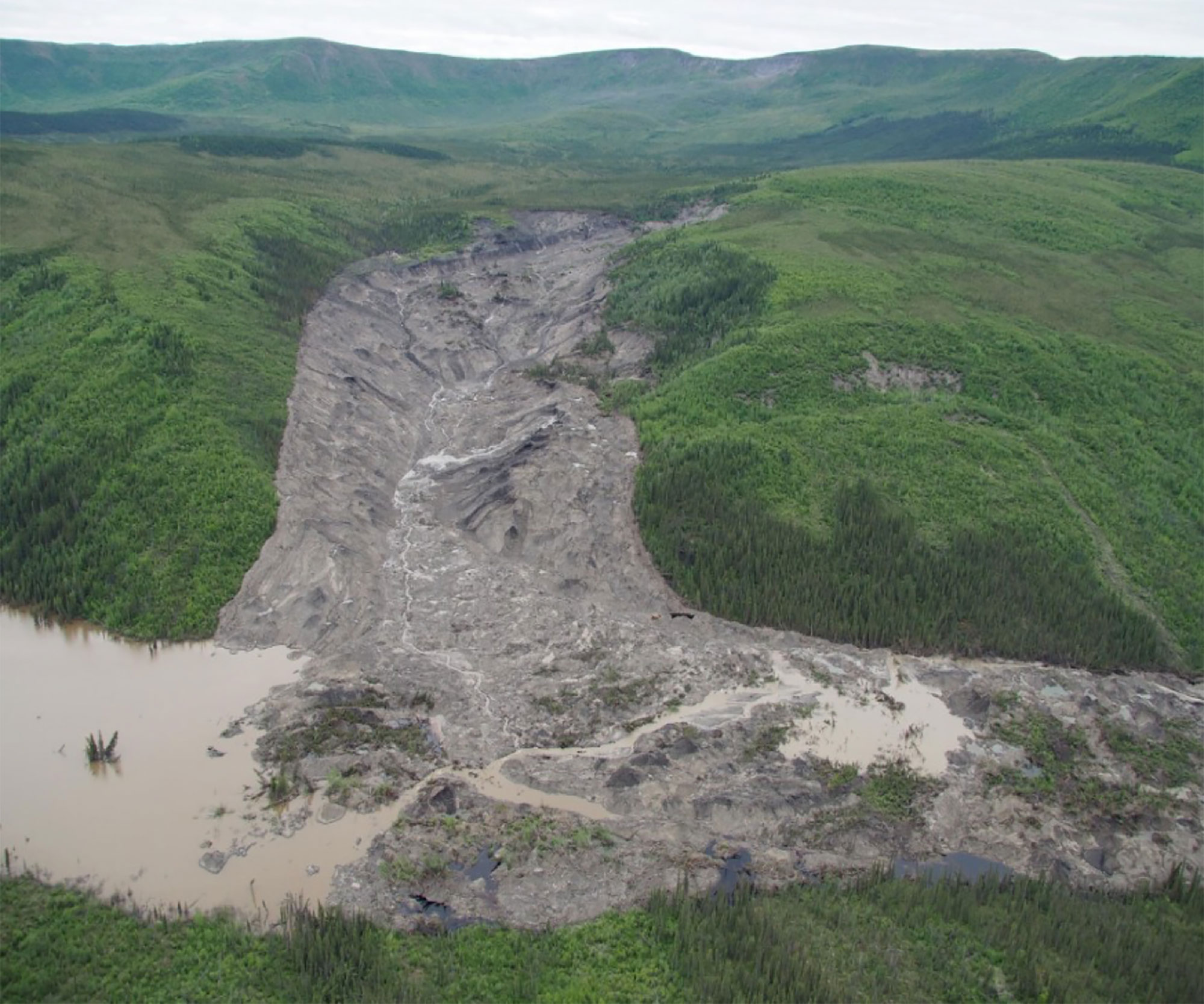 Image via Grist landscape showing damaged earth caused by permafrost detachment slides with tree covered hills on either side — Image via Grist