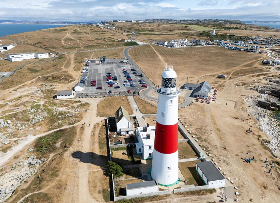 An aerial view shows Portland lighthouse next to a car park and a wide view of brown grass and vegetation, with the sea and a cloudy blue sky in the background, taken on Thursday.